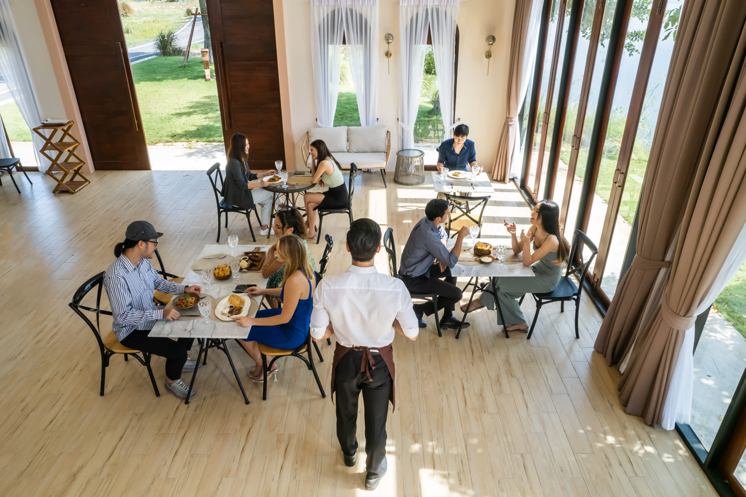 Caucasian waiter serving food to group of diverse customer in restaurant. Attractive male server service man work by delivering meal and drink to consumer at their table in dining room with happiness.